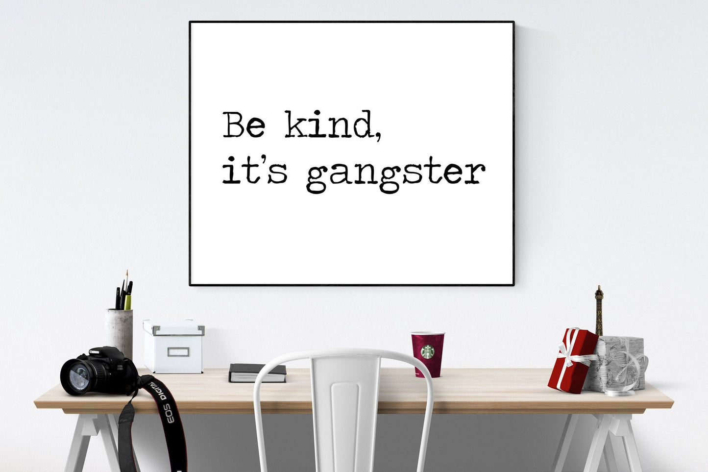 May include: A black and white framed print with the text 'Be kind, it's gangster' hanging on a white wall above a wooden desk with a white chair, a camera, a cup of coffee, and a small gift box.