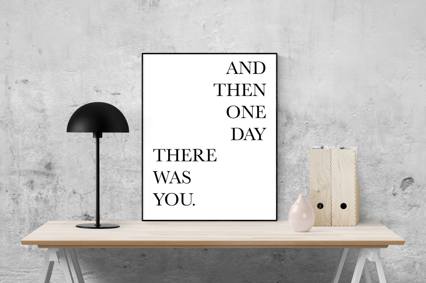 May include: A black and white poster with the text 'AND THEN ONE DAY THERE WAS YOU.' The poster is hanging on a gray wall above a wooden table with a black lamp and a pink vase.