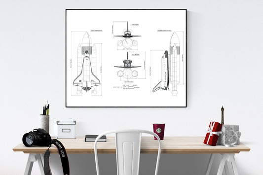 May include: A black and white line drawing of a space shuttle in three different views. The drawing is framed and hanging on a white wall above a desk with a camera, a cup of coffee, and a small Eiffel Tower statue.