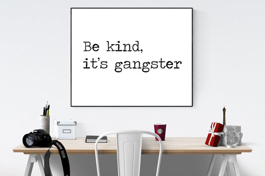 May include: A black and white framed print with the text &#39;Be kind, it&#39;s gangster&#39; hanging on a white wall above a wooden desk with a white chair, a camera, a cup of coffee, and a small gift box.