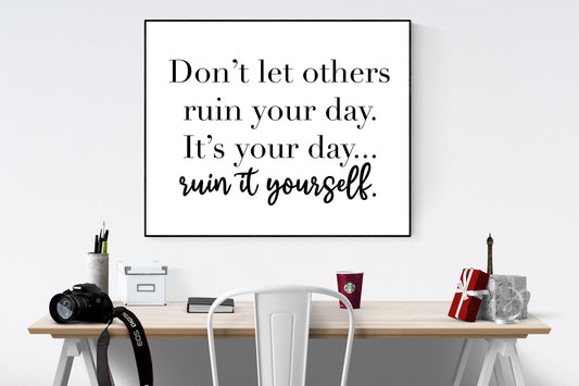 May include: A black and white framed motivational quote that reads &#39;Don&#39;t let others ruin your day. It&#39;s your day... ruin it yourself.&#39; The quote is hanging on a white wall above a wooden desk with a white chair, a camera, a cup of coffee, and a small gift box.