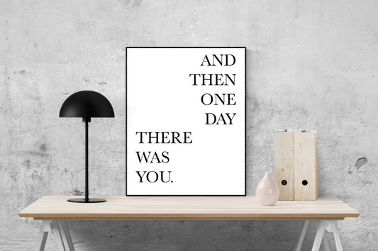 May include: A black and white poster with the text &#39;AND THEN ONE DAY THERE WAS YOU.&#39; The poster is hanging on a gray wall above a wooden table with a black lamp and a pink vase.