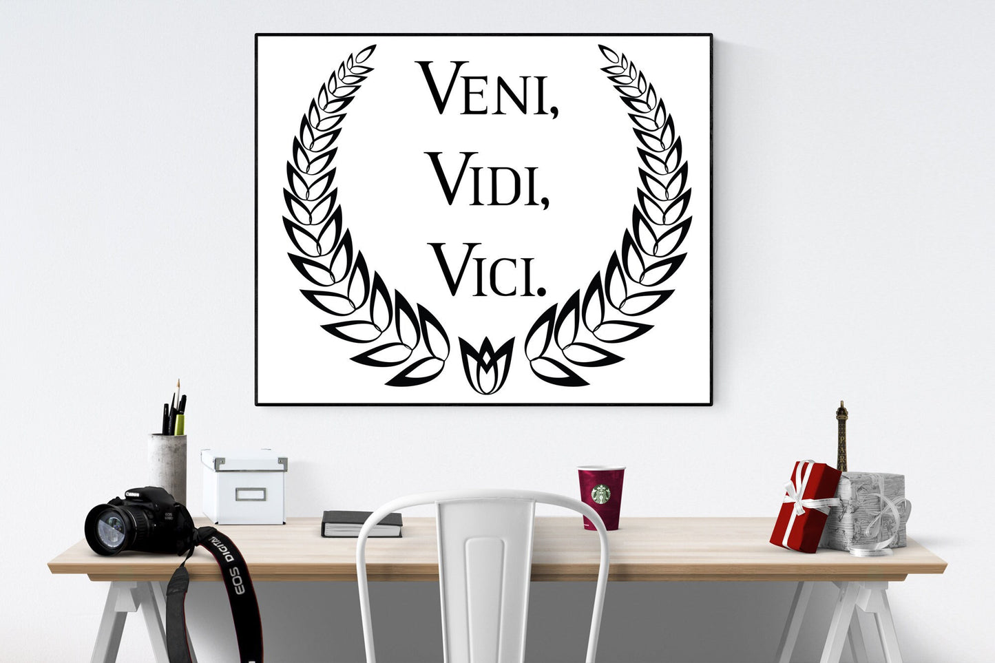 May include: A black and white print with a laurel wreath surrounding the text 'VENI, VIDI, VICI.' The print is framed and hanging on a white wall above a wooden desk with a white chair, a camera, a cup of coffee, and a small gift box.