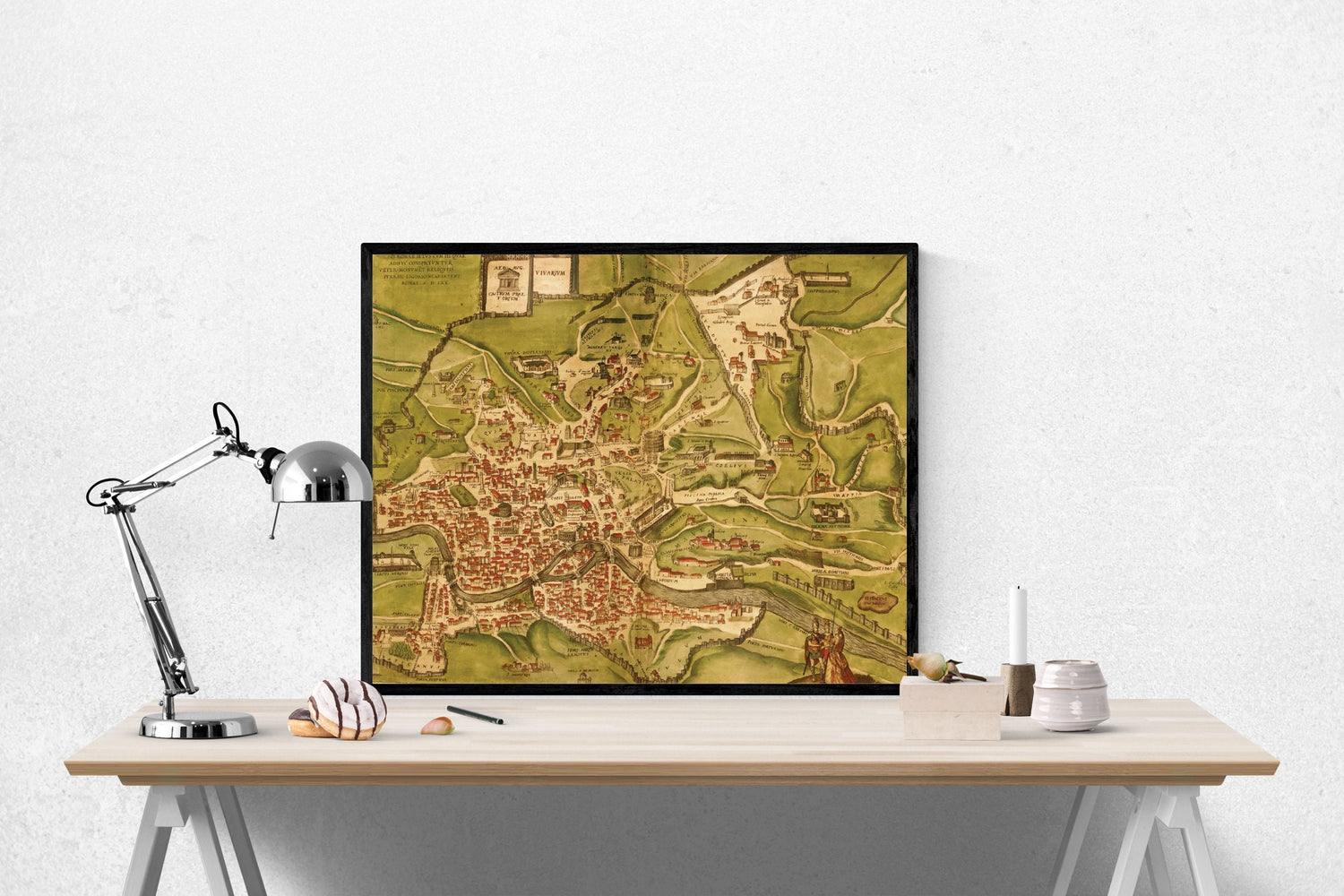 May include: A framed vintage map of a city with a green and brown color scheme. The map is displayed on a white desk with a silver lamp, a white donut, and a small white ceramic vase with a candle.