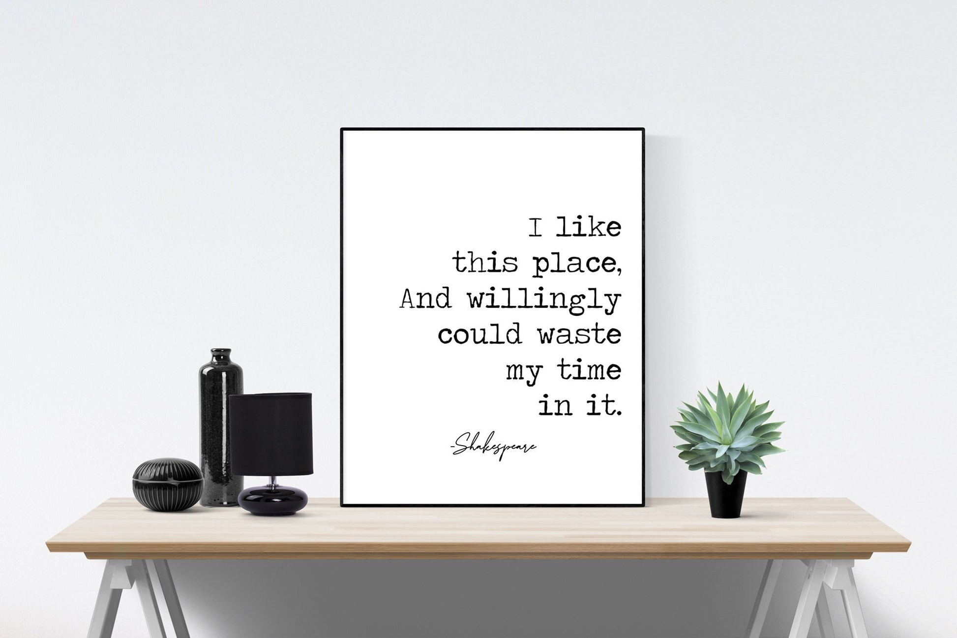 May include: A black and white typography print with the quote &#39;I like this place, And willingly could waste my time in it.&#39; with the signature &#39;Shakespeare&#39; below the quote. The print is framed and displayed on a wooden table with a black vase, a black lamp, and a small green succulent plant.