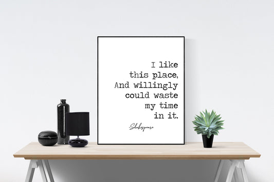 May include: A black and white typography print with the quote &#39;I like this place, And willingly could waste my time in it.&#39; with the signature &#39;Shakespeare&#39; below the quote. The print is framed and displayed on a wooden table with a black vase, a black lamp, and a small green succulent plant.