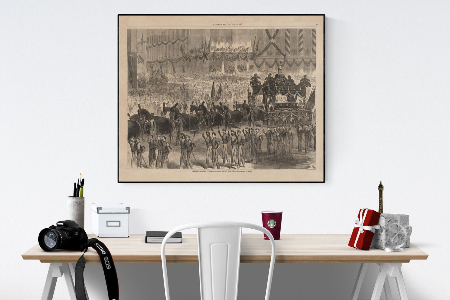 May include: A black and white framed print depicting a large-scale historical procession. The image shows a hearse drawn by horses, followed by uniformed soldiers and a large crowd of onlookers. The print has a vintage aesthetic and includes text at the bottom. The print is displayed on a light-colored wall above a desk with office supplies, a camera, and a coffee cup.