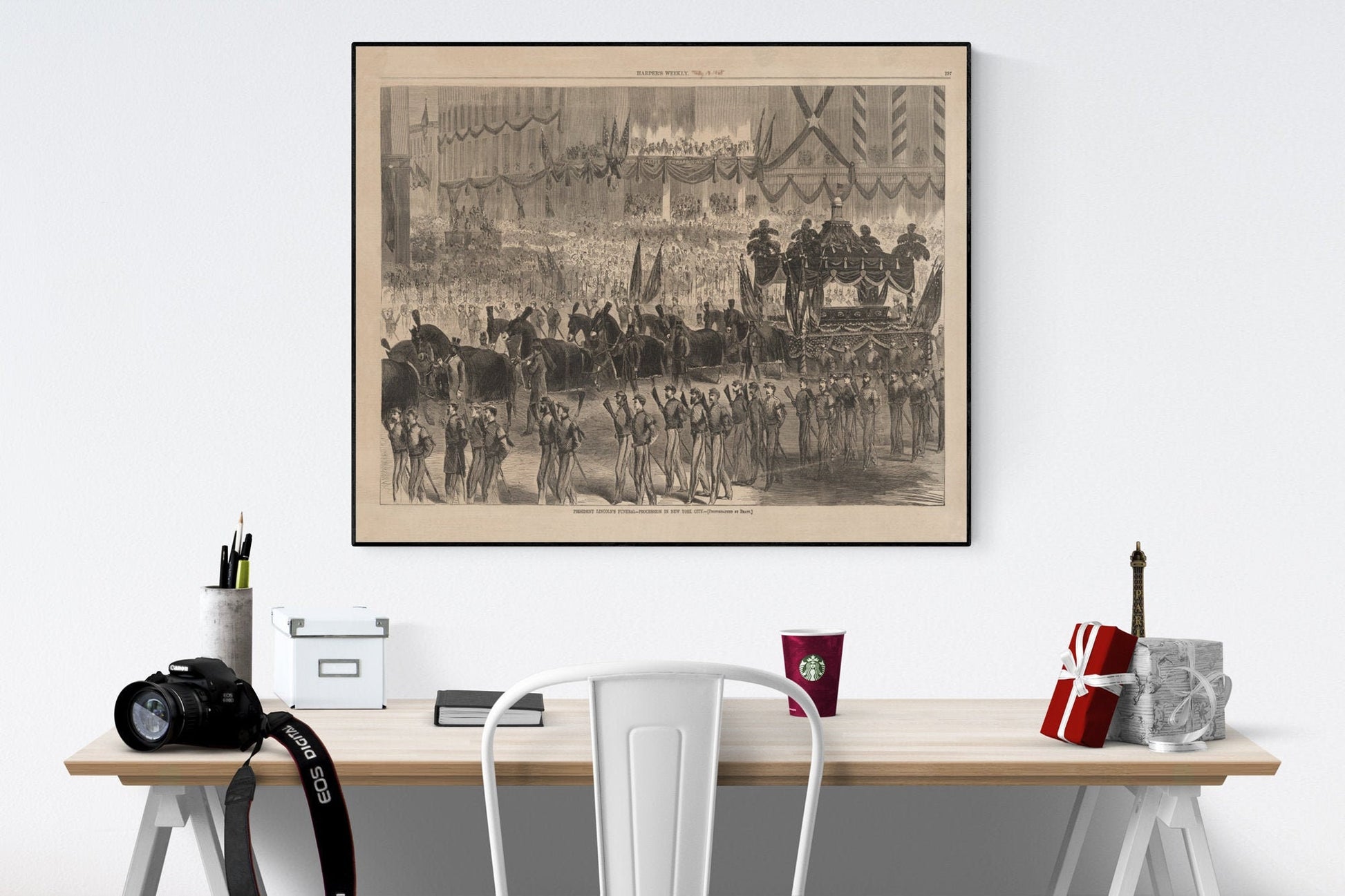 May include: A black and white framed print depicting a large-scale historical procession. The image shows a hearse drawn by horses, followed by uniformed soldiers and a large crowd of onlookers. The print has a vintage aesthetic and includes text at the bottom. The print is displayed on a light-colored wall above a desk with office supplies, a camera, and a coffee cup.