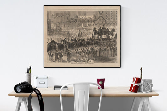 May include: A black and white framed print depicting a large-scale historical procession. The image shows a hearse drawn by horses, followed by uniformed soldiers and a large crowd of onlookers. The print has a vintage aesthetic and includes text at the bottom. The print is displayed on a light-colored wall above a desk with office supplies, a camera, and a coffee cup.