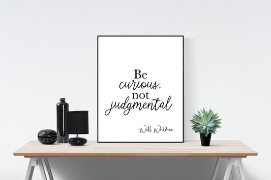 May include: A black and white typography print with the quote &#39;Be curious, not judgmental - Walt Whitman&#39; on a white background. The print is framed and displayed on a wooden table with a black vase, a black lamp, and a potted green succulent plant.
