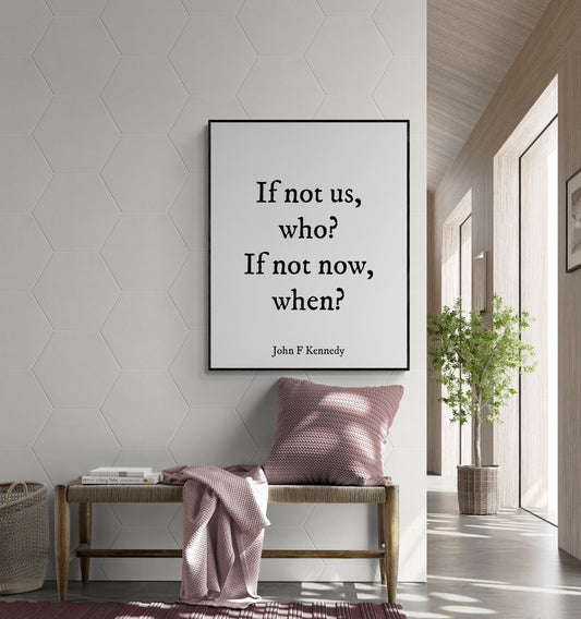 May include: A black and white framed print with the quote 'If not us, who? If not now, when?' by John F Kennedy. The print is hanging on a white wall with a hexagonal pattern. A wooden bench with a pink throw blanket and a pink pillow is in front of the print. A potted plant is in the background.