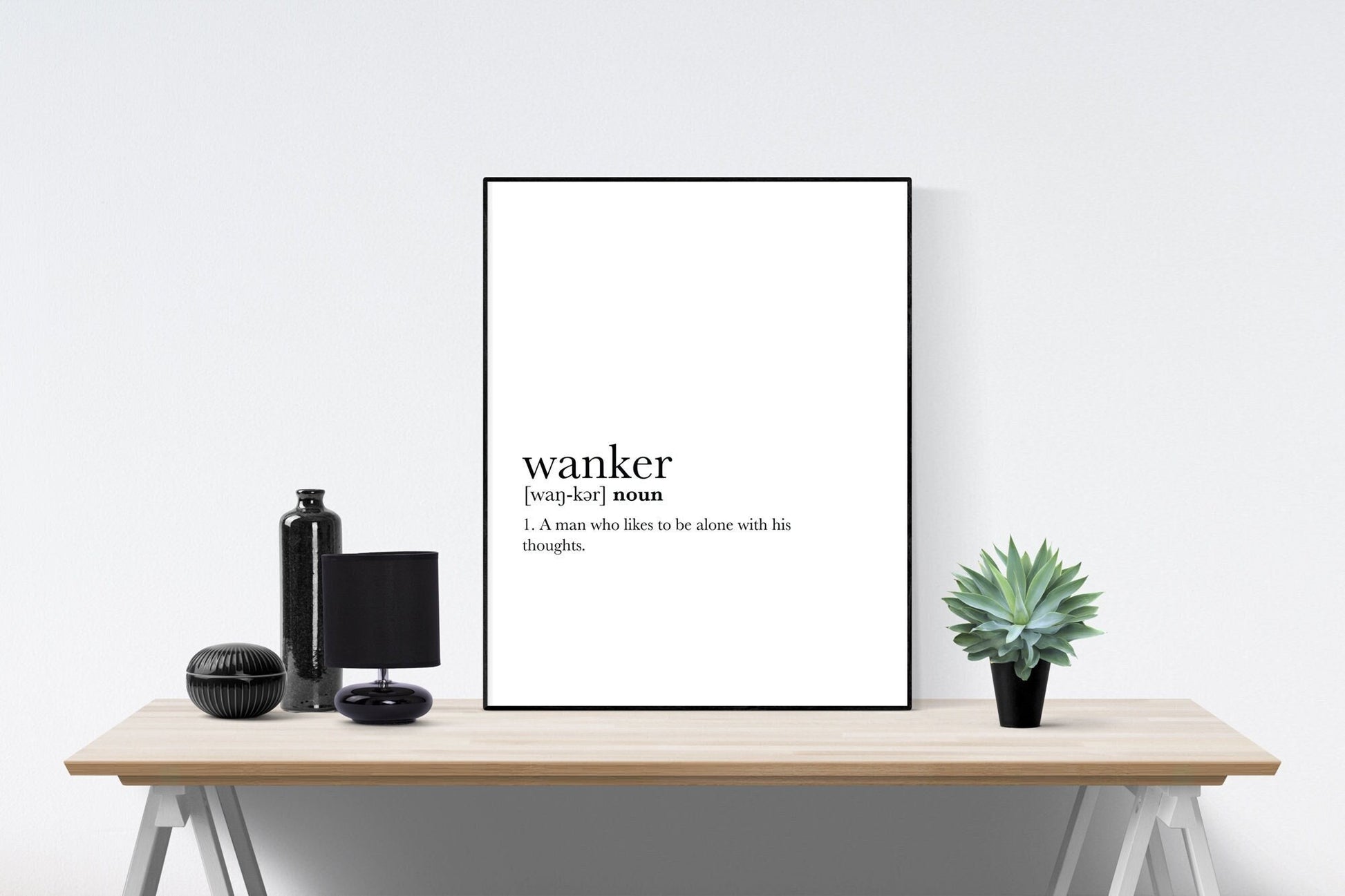 May include: A black and white print with the word &#39;wanker&#39; defined as &#39;A man who likes to be alone with his thoughts.&#39; The print is framed and displayed on a wooden table with a black lamp, a black bottle, and a potted green plant.