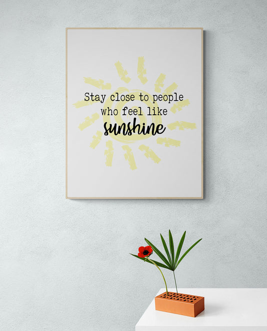 May include: A framed print with a yellow sun graphic and the text 'Stay close to people who feel like sunshine'. A red flower and green plant are in a brown brick vase on a white surface.