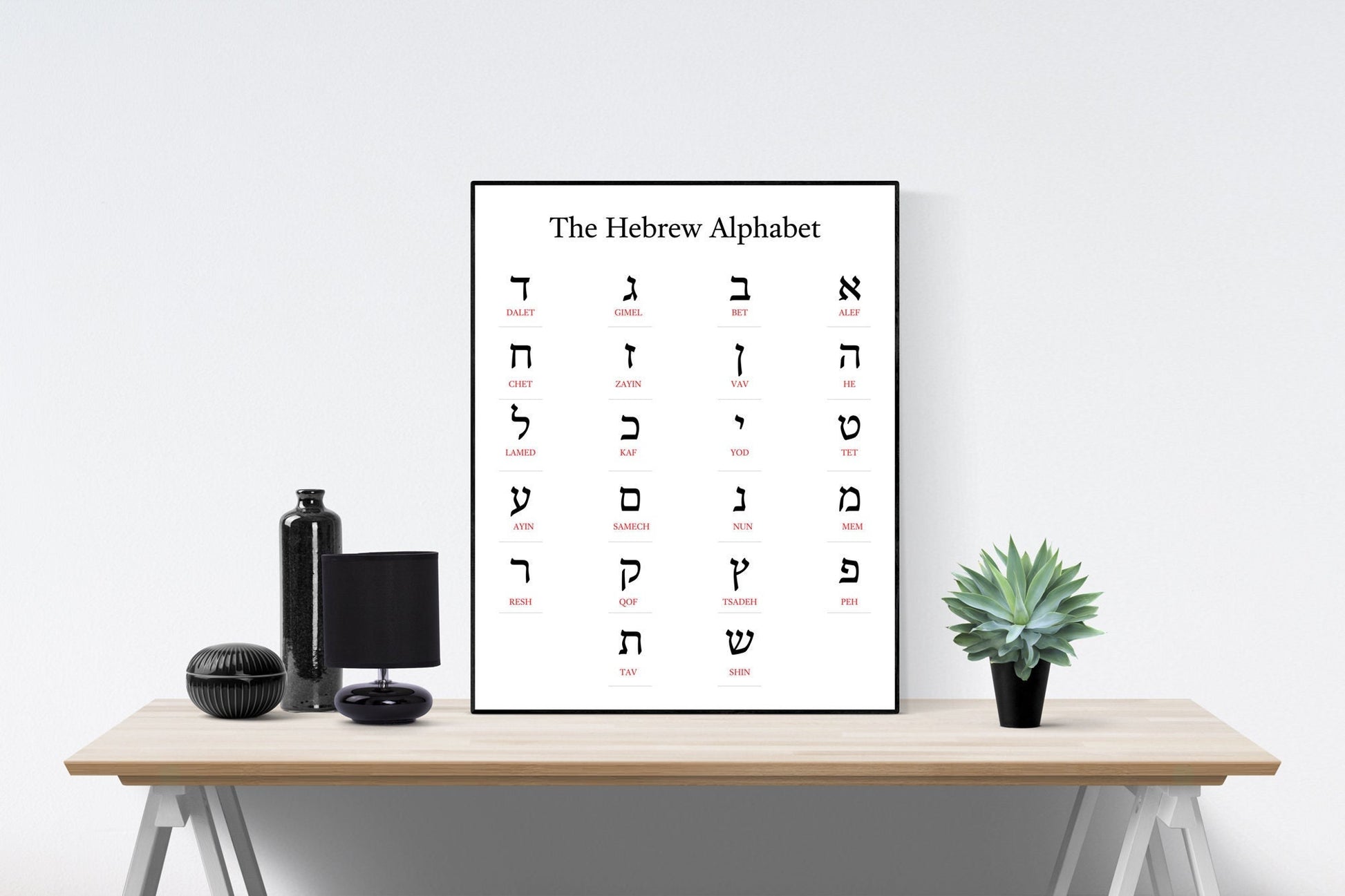 May include: A black and white poster with the Hebrew alphabet. The poster is framed and hanging on a white wall above a wooden table. The table has a black bottle, a black lamp, and a potted succulent plant on it.