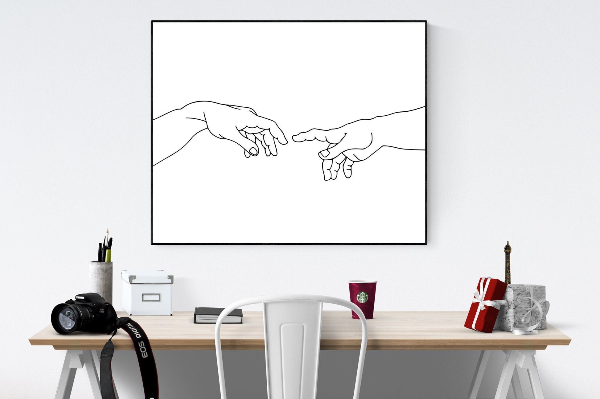 May include: A black and white line drawing of two hands reaching towards each other, framed in a black frame. The drawing is hanging on a white wall above a wooden desk with a white chair, a camera, a white box, a book, a cup, and a gift box on the desk.