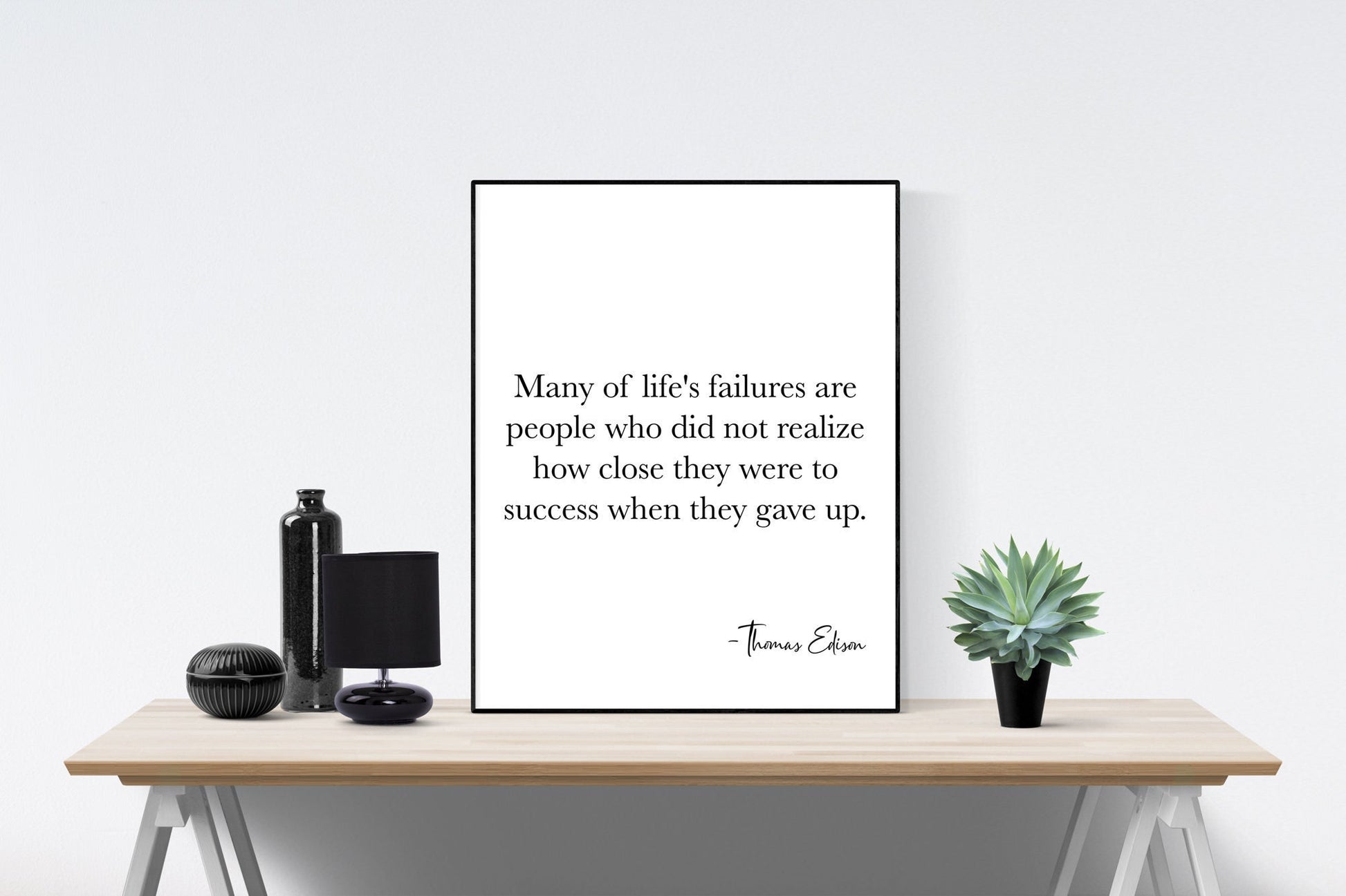 May include: A black and white motivational quote poster with the text 'Many of life's failures are people who did not realize how close they were to success when they gave up. -Thomas Edison' on a white background. The poster is framed in a black frame and is sitting on a wooden table with a black vase, a black lamp, and a potted succulent plant.