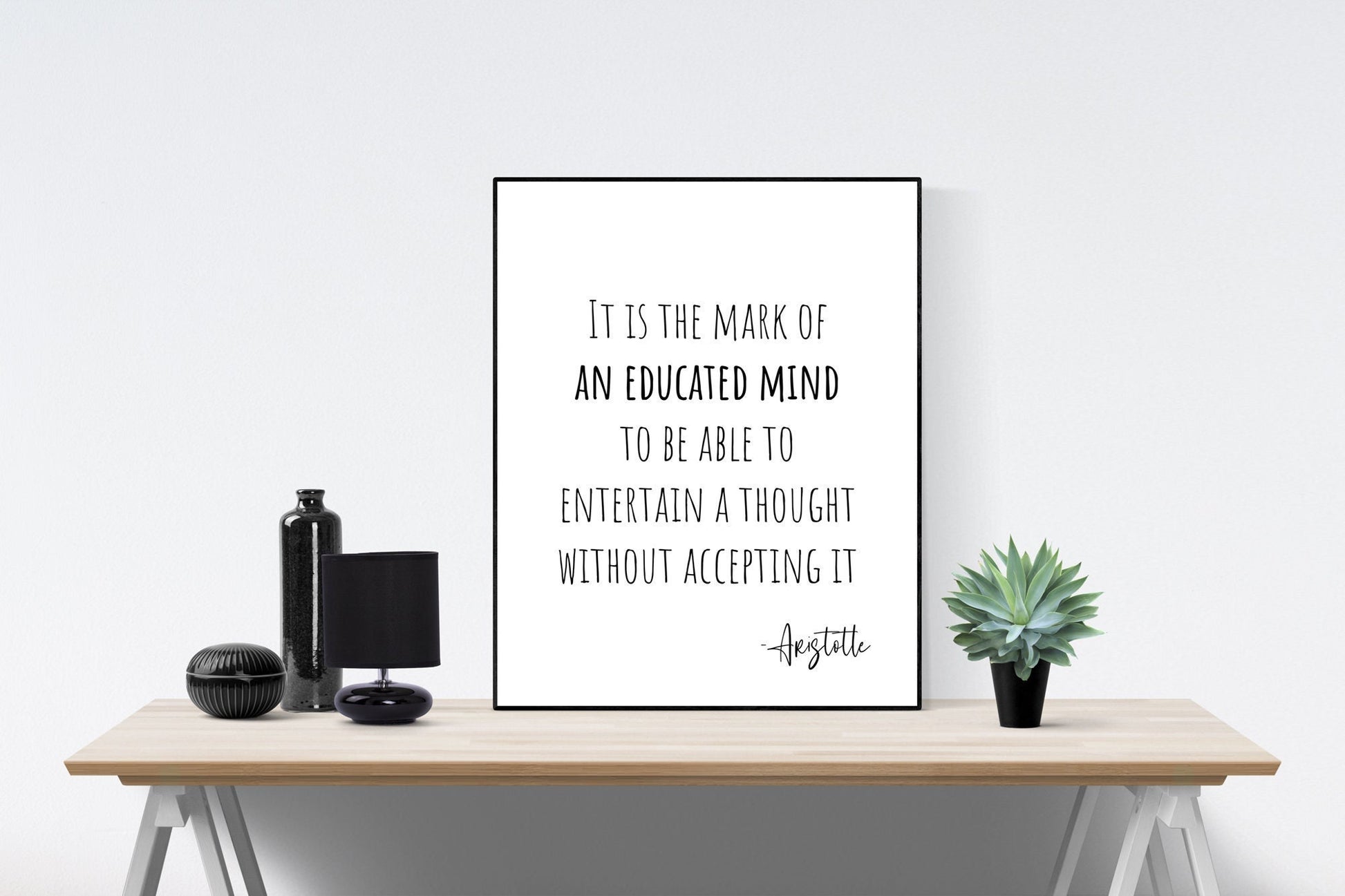 May include: A black and white framed print with the quote 'It is the mark of an educated mind to be able to entertain a thought without accepting it - Aristotle' on a white background. The print is sitting on a wooden table with a black vase, a black lamp, and a green succulent plant.