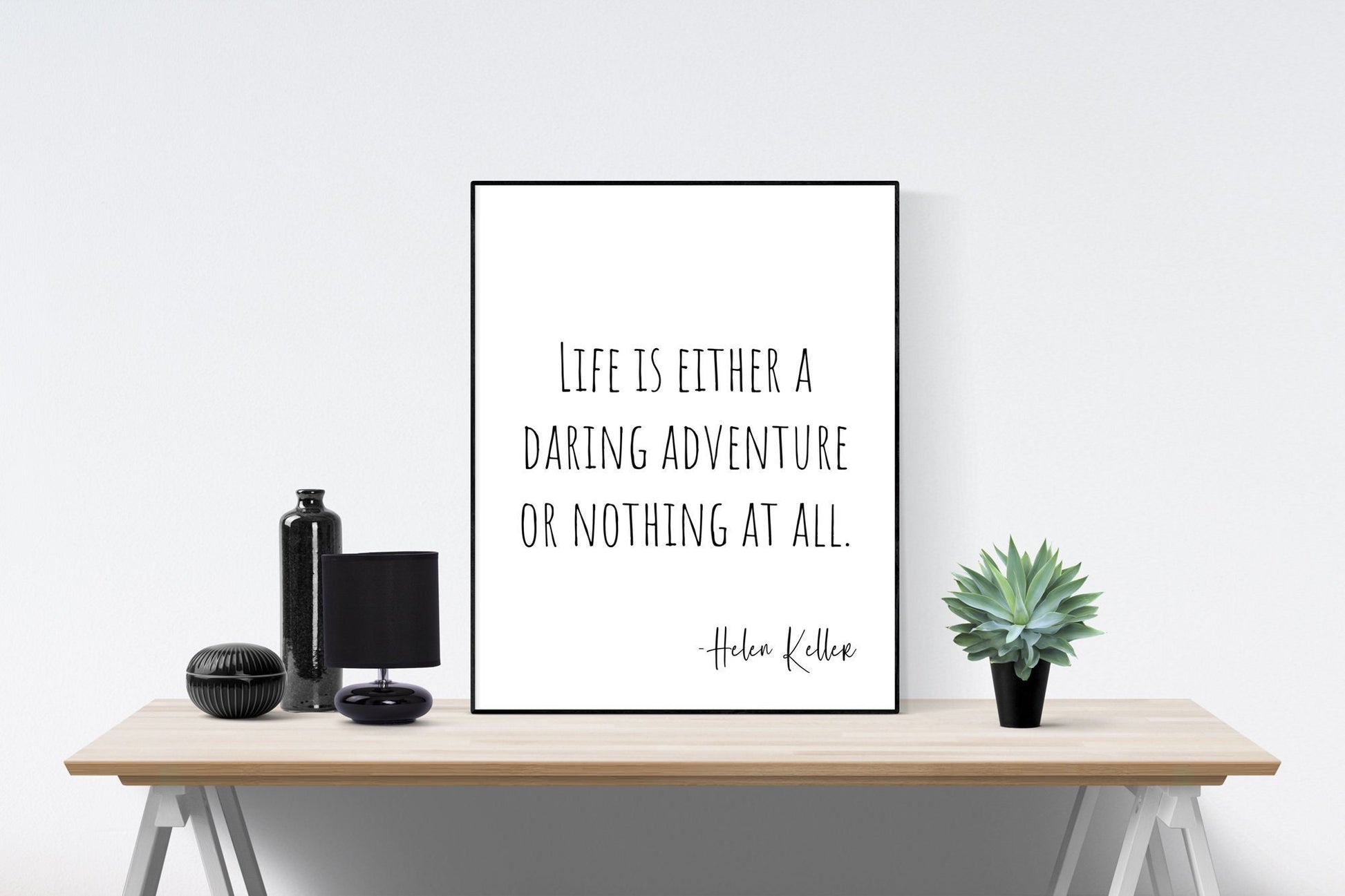 May include: A black and white framed print with the quote 'Life is either a daring adventure or nothing at all. -Helen Keller' on a white background. The print is sitting on a wooden table with a black bottle, a black lamp, and a potted green plant.