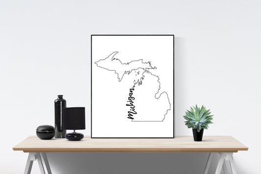May include: A black and white line art print of the state of Michigan with the word 'Michigan' written vertically along the state's border. The print is framed and displayed on a wooden table with a black vase, a black lamp, and a green succulent plant.