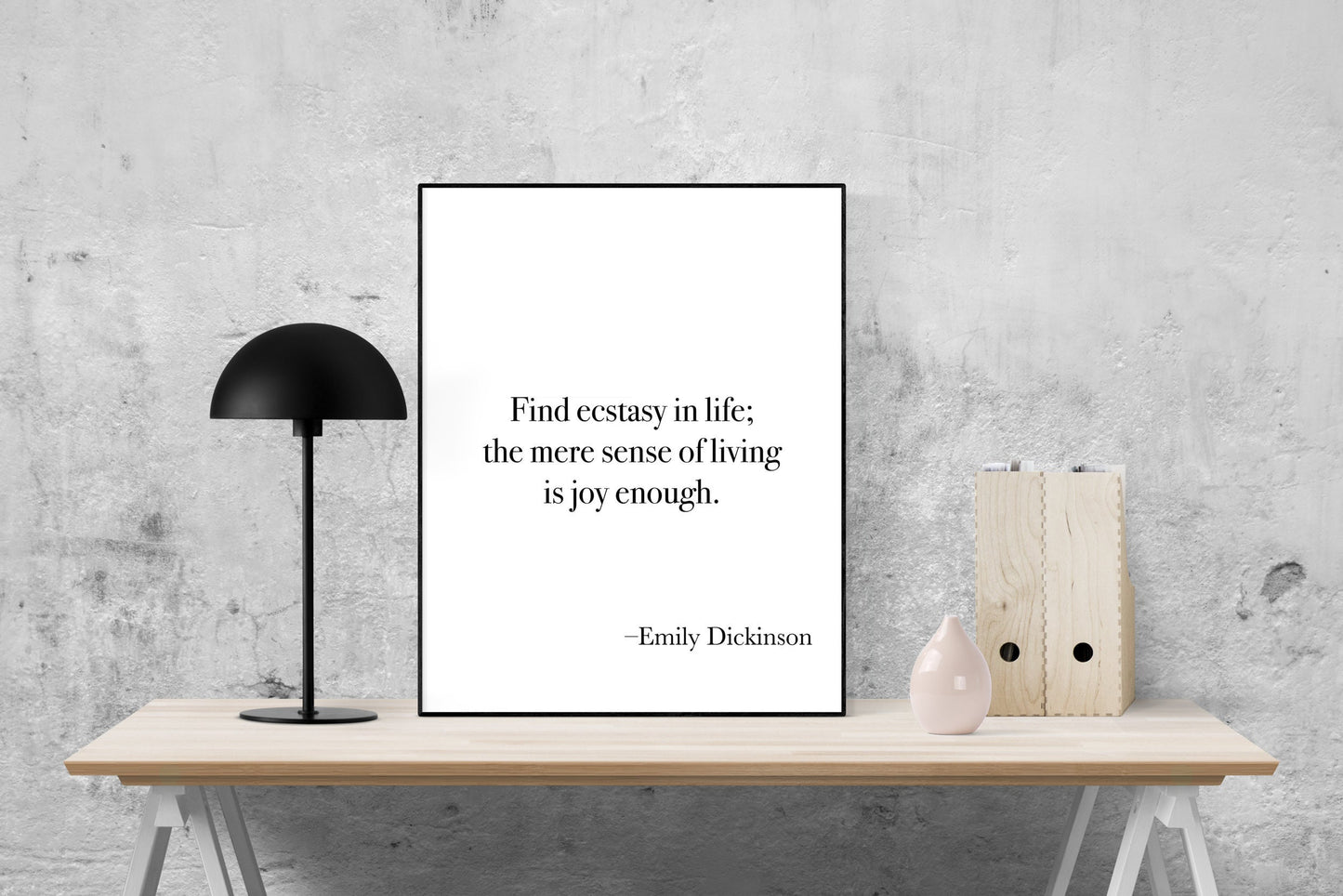 May include: A black and white printable quote poster with the text 'Find ecstasy in life; the mere sense of living is joy enough. -Emily Dickinson' displayed on a white background. The poster is framed with a black frame and is sitting on a light brown wooden table.