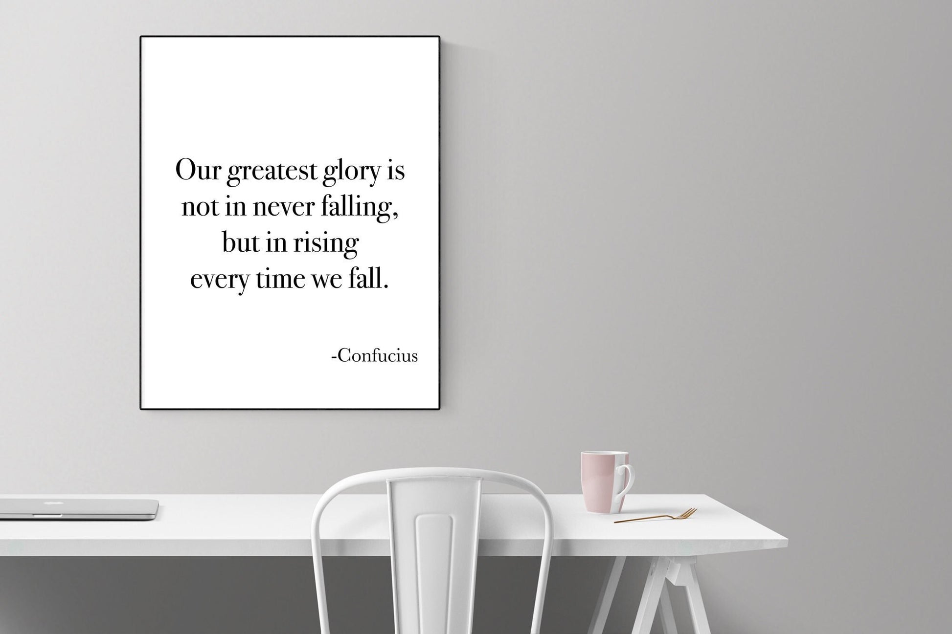 May include: A black and white framed print with the quote 'Our greatest glory is not in never falling, but in rising every time we fall. -Confucius' hanging on a light gray wall above a white table with a white chair and a pink mug on the table.