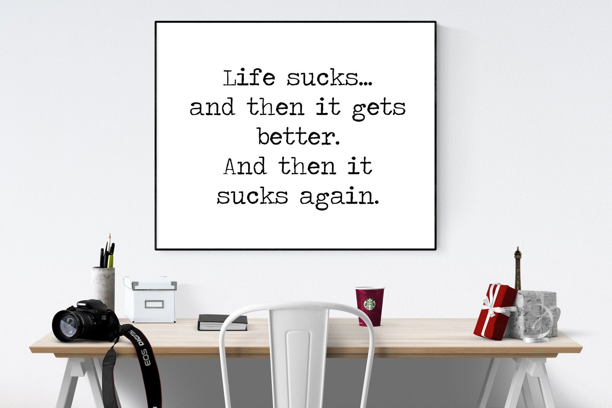 May include: A black and white framed print with the text 'Life sucks... and then it gets better. And then it sucks again.' The print is hanging on a white wall above a wooden desk with a white chair, a camera, a red cup, a small Eiffel Tower figurine, and a wrapped gift box.