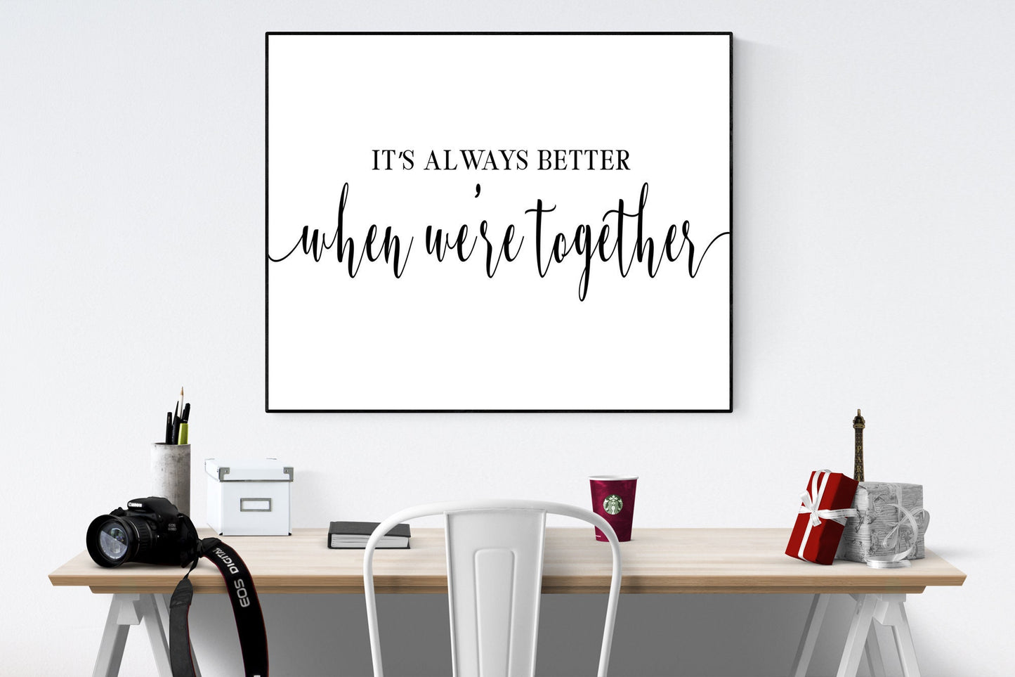 May include: A black and white framed print with the text 'It's always better when we're together' on a white background. The print is hanging on a white wall above a wooden desk with a white chair, a camera, a cup of coffee, and a small gift box.