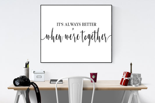 May include: A black and white framed print with the text 'It's always better when we're together' on a white background. The print is hanging on a white wall above a wooden desk with a white chair, a camera, a cup of coffee, and a small gift box.