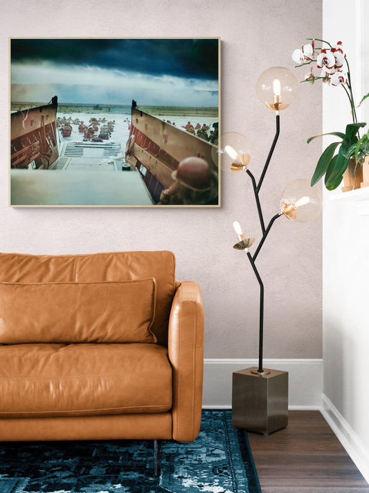 May include: A brown leather sofa with a black floor lamp with multiple round glass globes. The lamp is on a dark gray square base. A blue rug is in front of the sofa. A framed picture of a black and white photo of a landing craft with soldiers is on the wall behind the sofa.