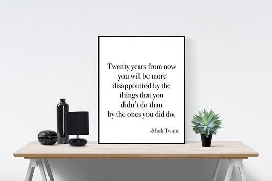 May include: A black and white framed print with a quote by Mark Twain that reads: 'Twenty years from now you will be more disappointed by the things that you didn't do than by the ones you did do.' The print is sitting on a wooden table with a black vase, a black lamp, and a small green plant.