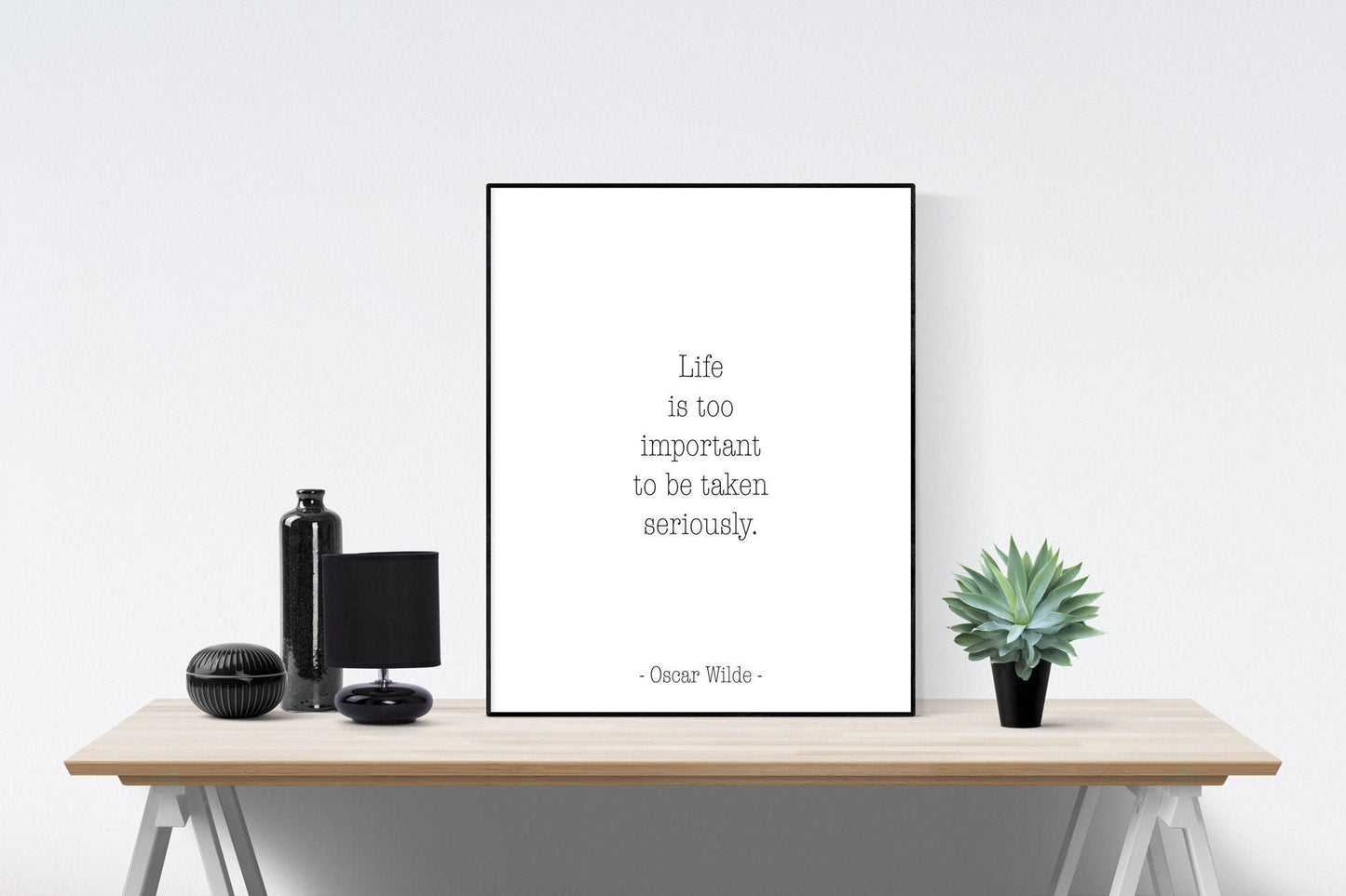 May include: A black and white framed print with the quote 'Life is too important to be taken seriously.' - Oscar Wilde. The print is sitting on a wooden table with a black bottle, a black lamp, and a green succulent plant.