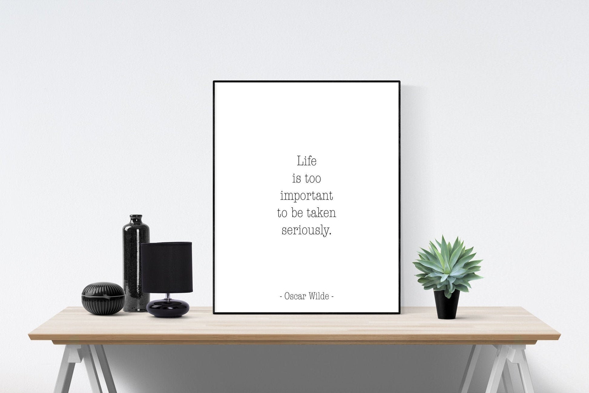 May include: A black and white framed print with the quote 'Life is too important to be taken seriously.' - Oscar Wilde. The print is sitting on a wooden table with a black bottle, a black lamp, and a green succulent plant.