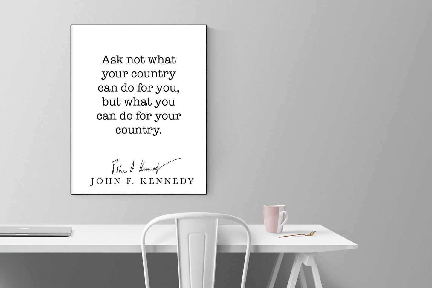 May include: A black and white framed print with the quote 'Ask not what your country can do for you, but what you can do for your country.'  The quote is attributed to John F. Kennedy with his signature below. The print is hanging on a gray wall above a white desk with a white chair and a pink mug on the desk.