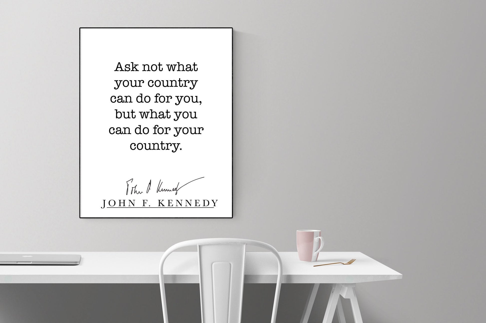 May include: A black and white framed print with the quote 'Ask not what your country can do for you, but what you can do for your country.'  The quote is attributed to John F. Kennedy with his signature below. The print is hanging on a gray wall above a white desk with a white chair and a pink mug on the desk.