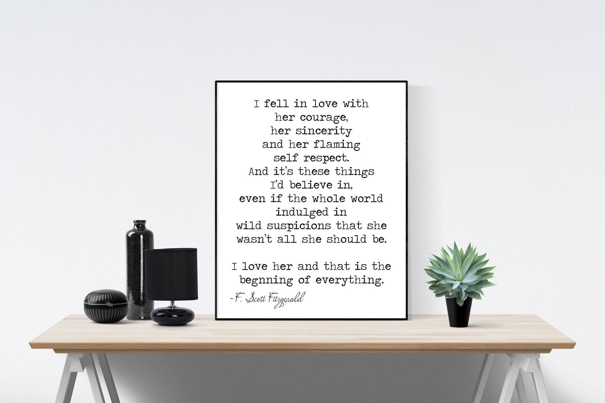 May include: A black and white framed print with a quote by F. Scott Fitzgerald that reads: 'I fell in love with her courage, her sincerity and her flaming self respect. And it's these things I'd believe in, even if the whole world indulged in wild suspicions that she wasn't all she should be. I love her and that is the beginning of everything. -F. Scott Fitzgerald' The print is sitting on a wooden table with a black lamp and a small potted succulent plant.