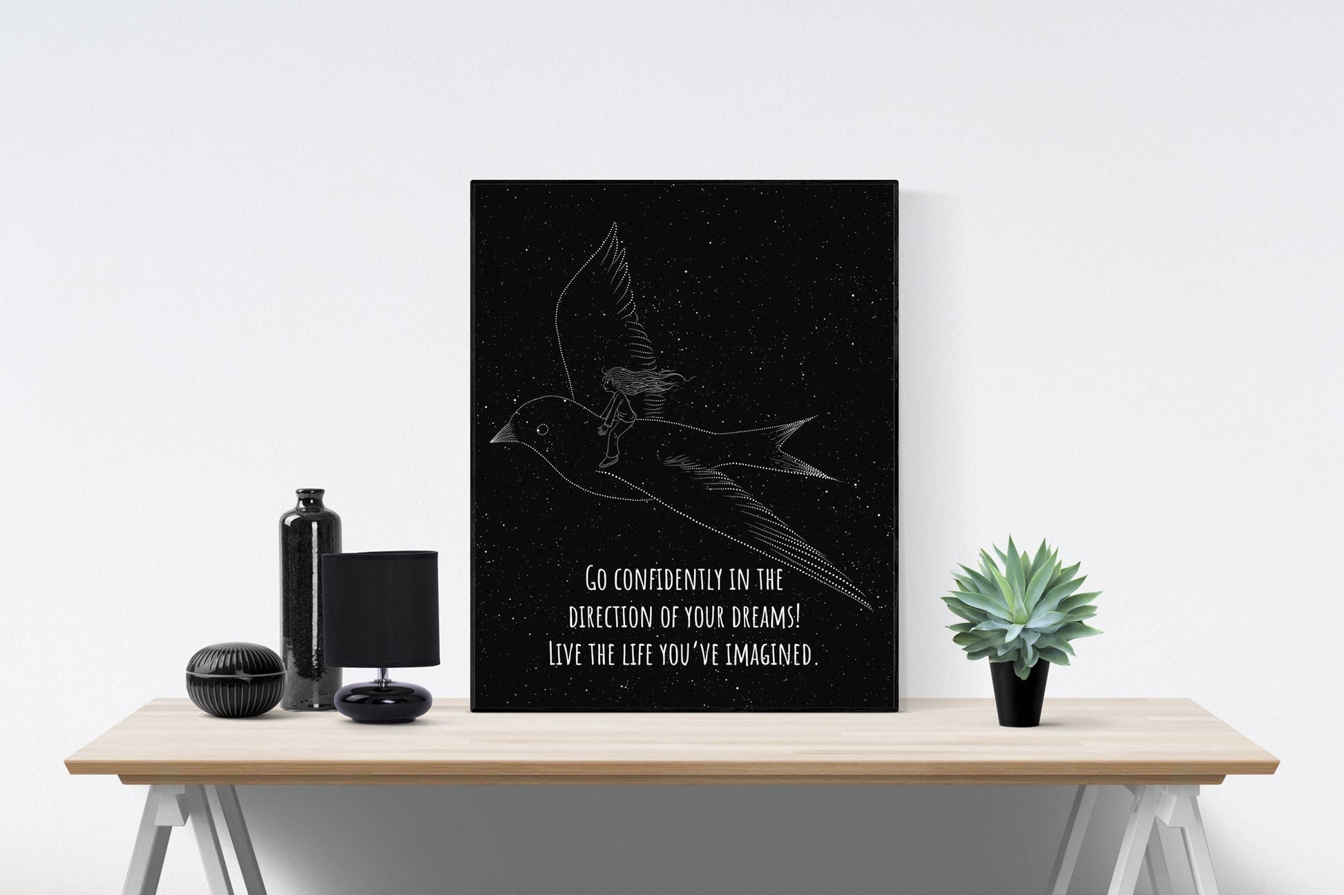May include: A black and white poster with a line drawing of a person flying on a bird. The poster has the text 'GO CONFIDENTLY IN THE DIRECTION OF YOUR DREAMS! LIVE THE LIFE YOU'VE IMAGINED.' The poster is displayed on a wooden table with a black vase, a black lamp, and a green plant.