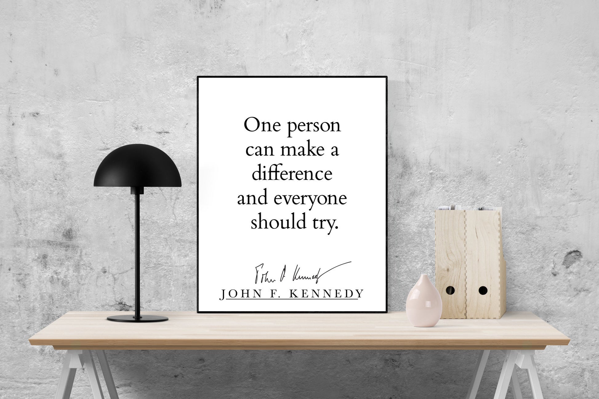 May include: A black and white typography print with the quote 'One person can make a difference and everyone should try.' The quote is attributed to John F. Kennedy with his signature below. The print is framed and displayed on a wooden table with a black lamp and a pink ceramic vase.