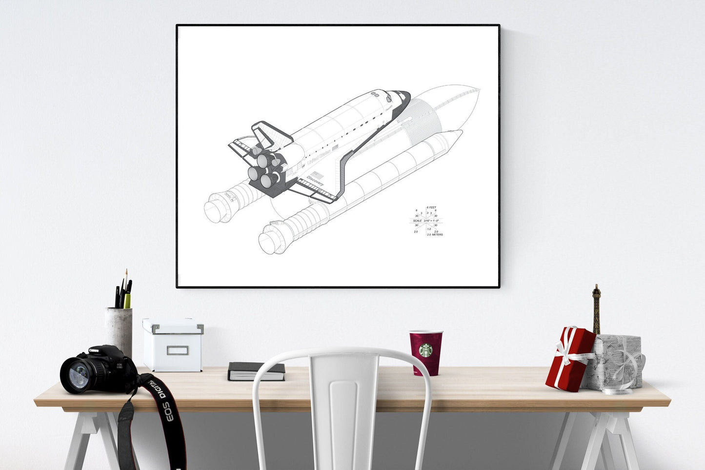 May include: A black and white line drawing of a space shuttle with its booster rockets. The drawing is framed and hanging on a white wall above a wooden desk with a camera, a cup of coffee, a book, and a small Eiffel Tower figurine.