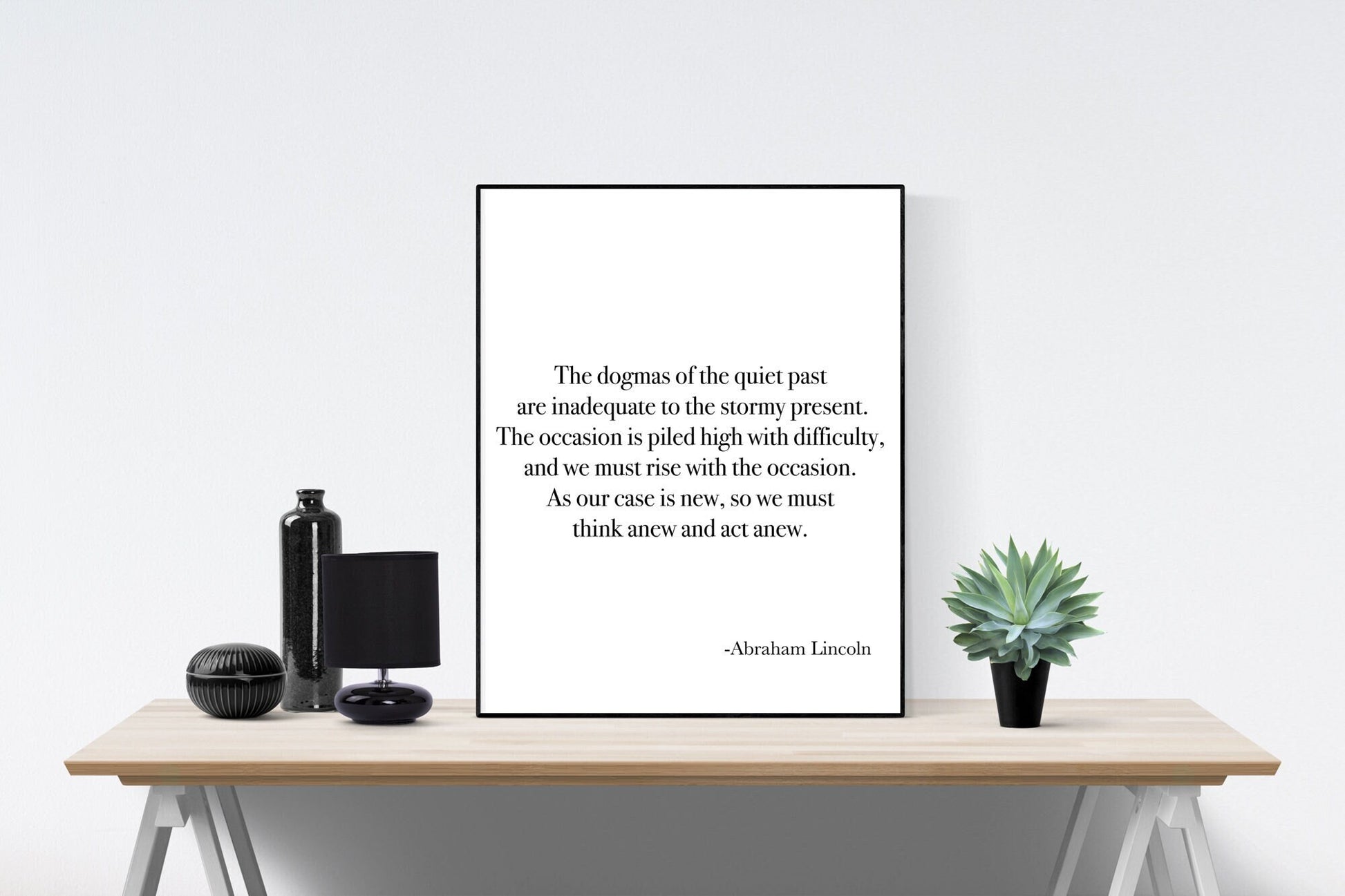 May include: A black and white framed print with a quote by Abraham Lincoln: 'The dogmas of the quiet past are inadequate to the stormy present. The occasion is piled high with difficulty, and we must rise with the occasion. As our case is new, so we must think anew and act anew.' The print is sitting on a wooden table with a black bottle and a black lamp on the left and a green succulent plant in a black pot on the right.