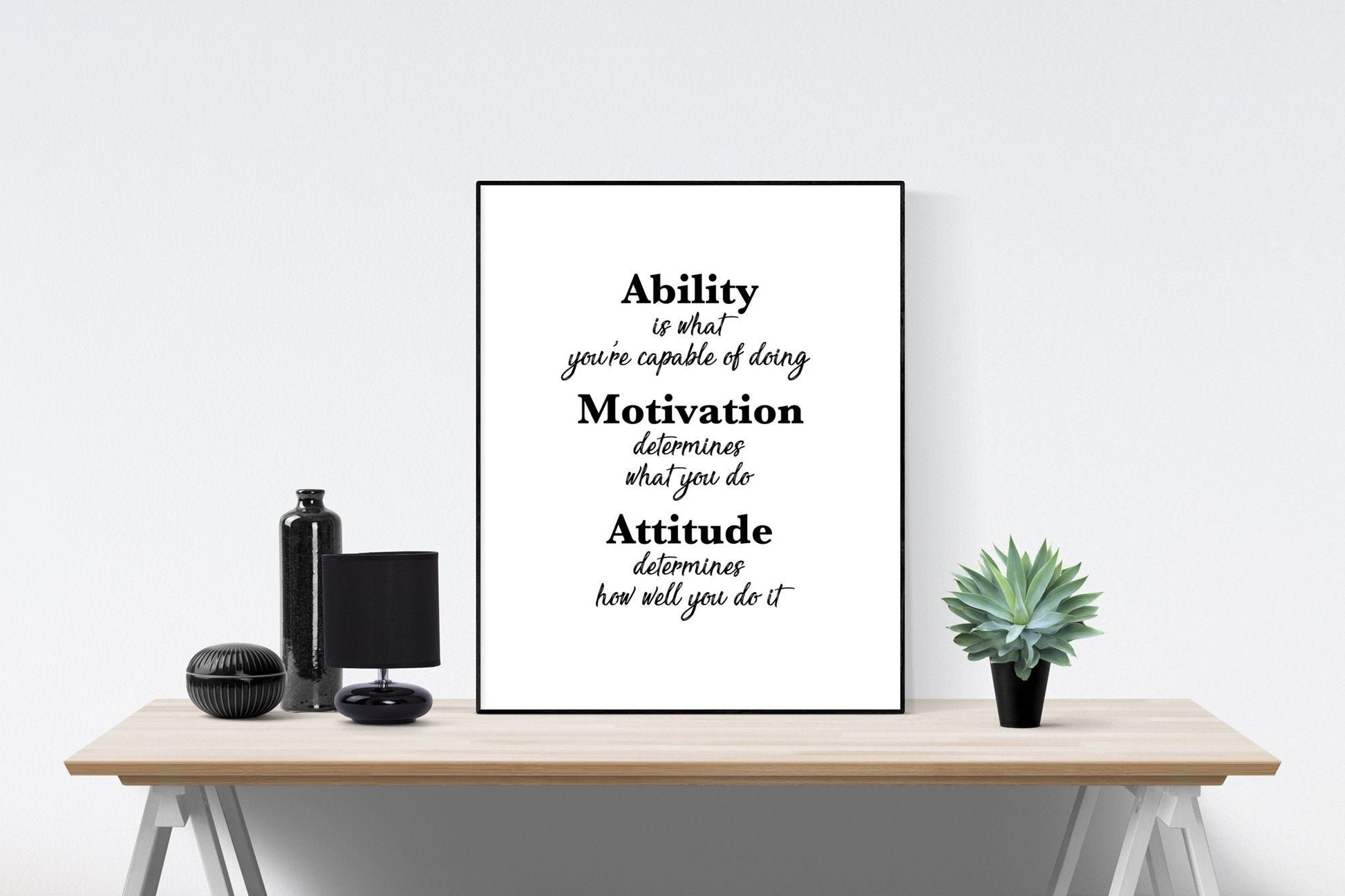 May include: A black and white motivational quote poster with the text 'Ability is what you're capable of doing. Motivation determines what you do. Attitude determines how well you do it.' The poster is framed and hanging on a white wall above a wooden table with a black lamp, a black vase, and a potted green plant.