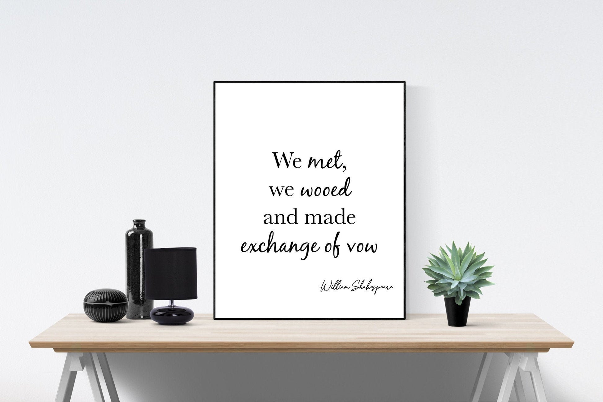 May include: A black and white framed print with the quote 'We met, we wooed and made exchange of vow' by William Shakespeare. The print is sitting on a wooden table with a black lamp, a black bottle, and a potted succulent plant.