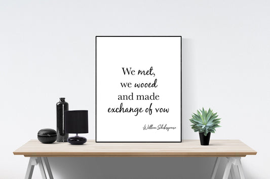 May include: A black and white framed print with the quote 'We met, we wooed and made exchange of vow' by William Shakespeare. The print is sitting on a wooden table with a black lamp, a black bottle, and a potted succulent plant.