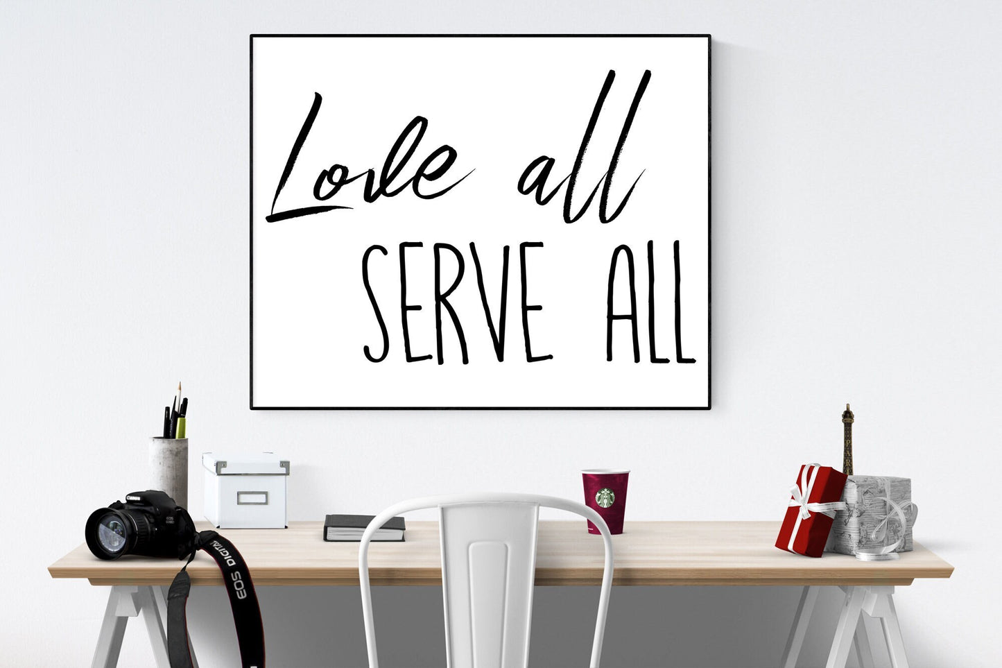 May include: A black and white framed print with the text 'Love all serve all' hanging on a white wall above a wooden desk with a white chair, a camera, a cup of coffee, and a gift box.