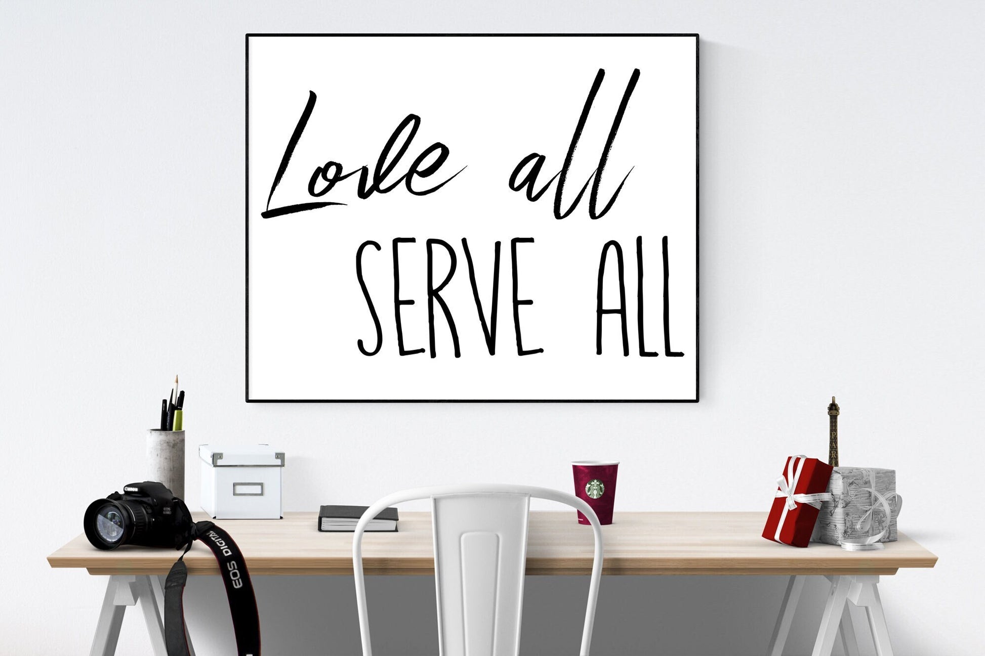 May include: A black and white framed print with the text 'Love all serve all' hanging on a white wall above a wooden desk with a white chair, a camera, a cup of coffee, and a gift box.