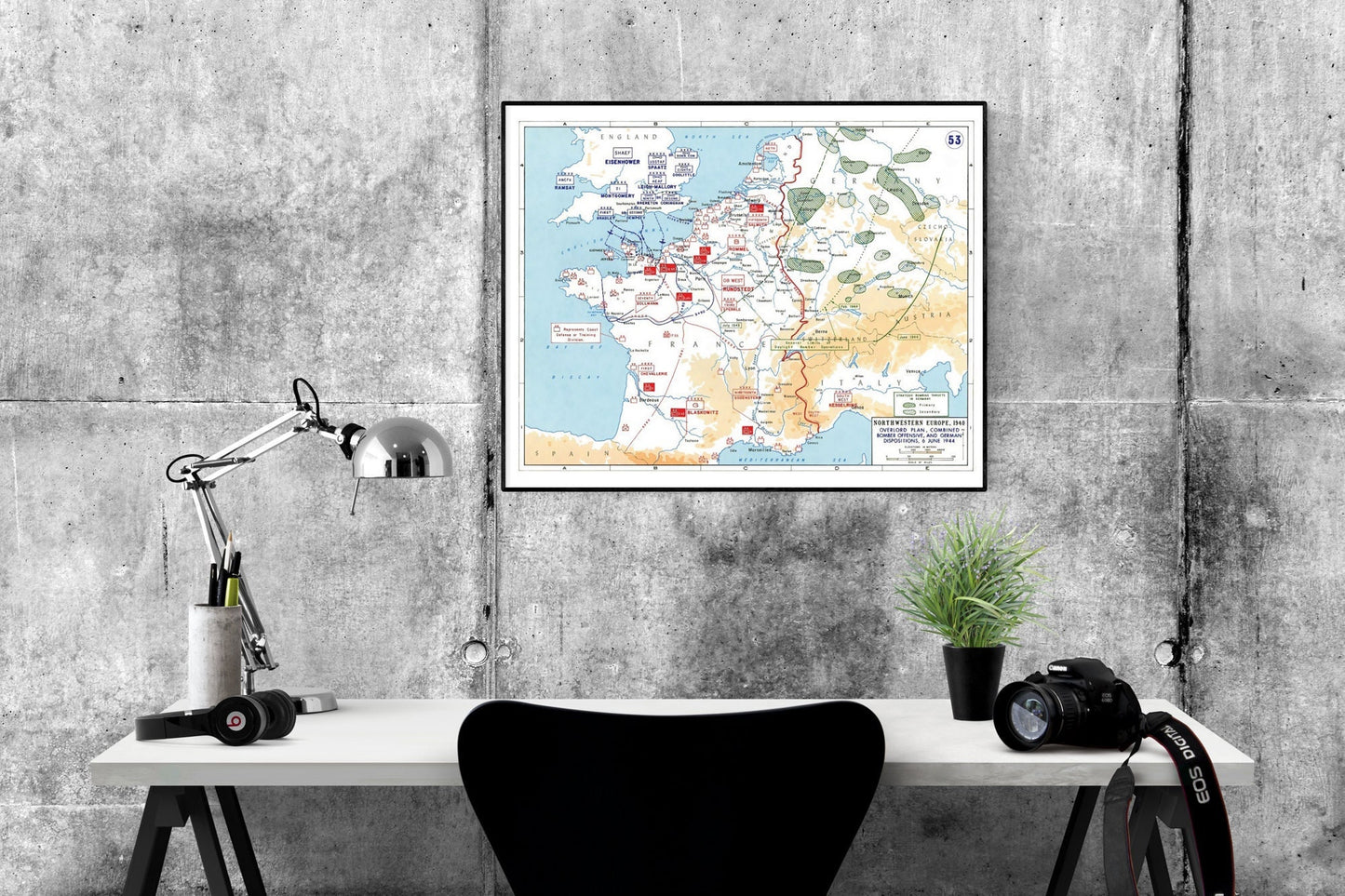 May include: A black and white map of France with red markings. The map is framed and hanging on a gray concrete wall. The map is titled 'WESTERN EUROPE 1944' and shows the Allied advance from Normandy to the German border. A desk with a lamp, headphones, a camera, and a plant is in the foreground.