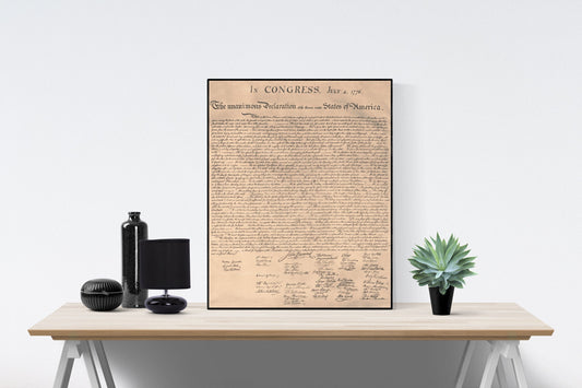 May include: A framed black and white print of the Declaration of Independence. The print is on a light brown background and has the text 'In CONGRESS, July 4, 1776. The unanimous Declaration of the thirteen united States of America.' The print is on a wooden table with a black vase, a black lamp, and a green plant.