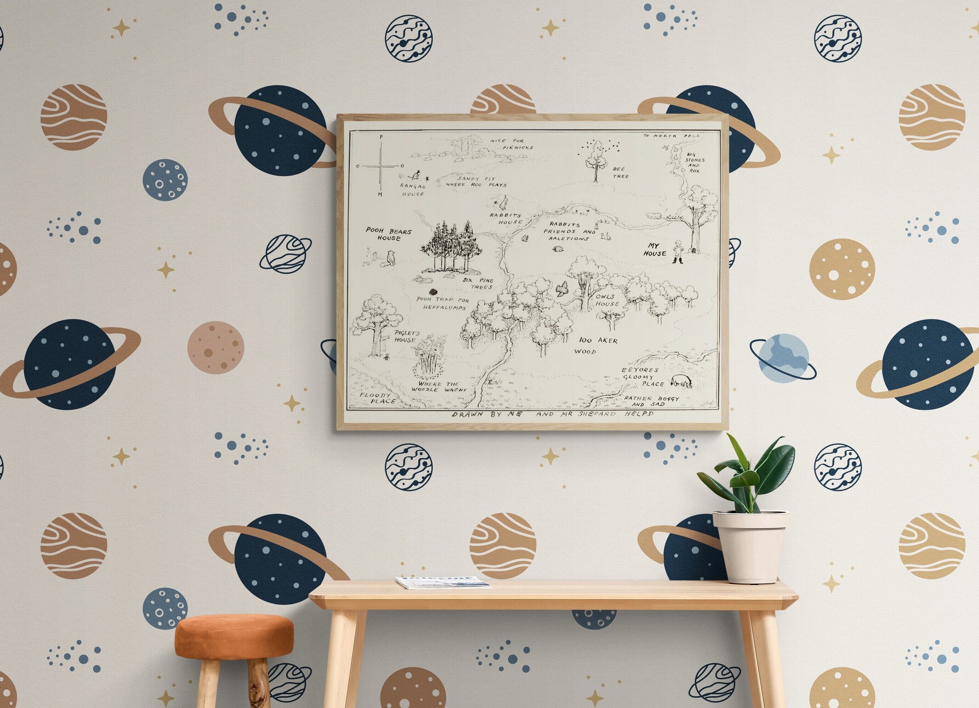 May include: A white wall with a blue and brown space themed wallpaper. The wallpaper features planets with rings, stars, and small dots. A framed map of a forest is hanging on the wall. A wooden table with a brown stool is in front of the wall. A potted plant is on the table.
