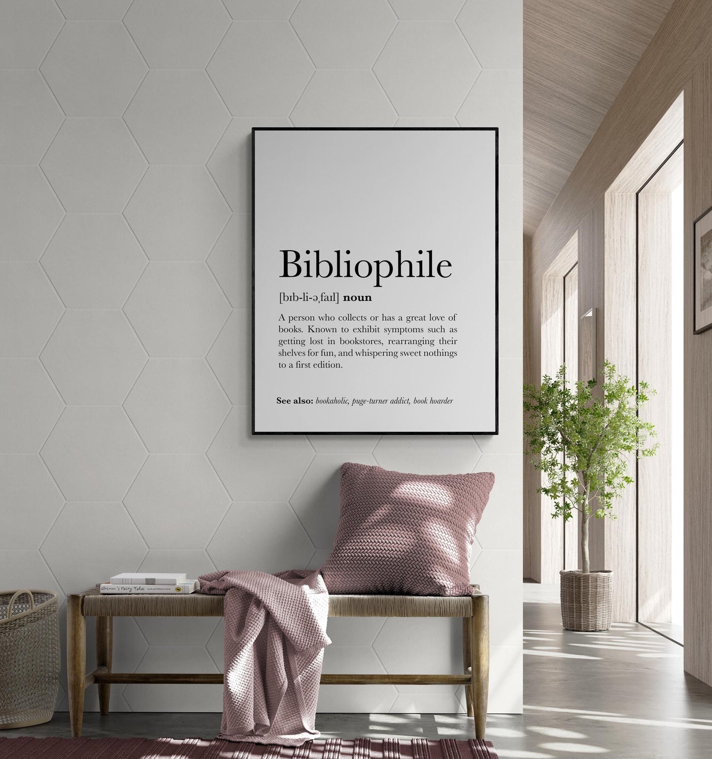 May include: Black and white definition print of the word 'Bibliophile' on a white background. The definition describes a bibliophile as someone who collects books or has a great love of books, exhibiting symptoms such as getting lost in bookstores, rearranging shelves for fun, and whispering sweet nothings to a first edition.  The print is displayed in a modern hallway setting with a bench and throw blanket in dusty rose, and a green plant in a woven basket.