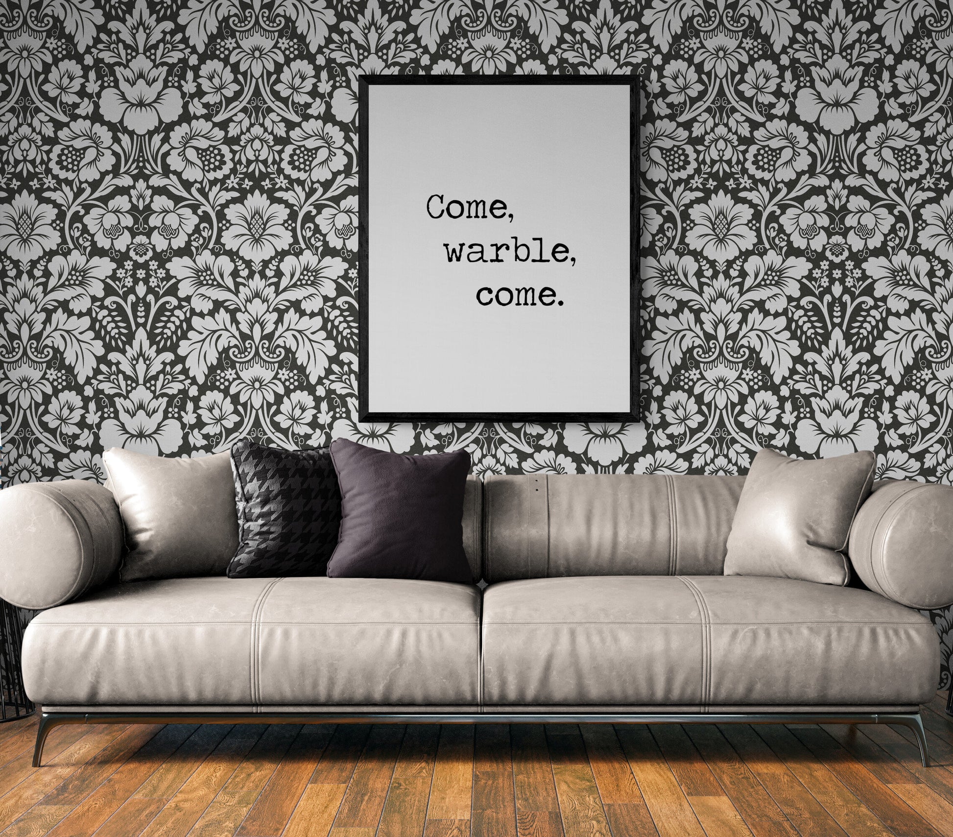 May include: A gray couch with black pillows in front of a black and white floral wallpaper. A black framed picture with the text 'Come, warble, come.' is hanging on the wall above the couch.